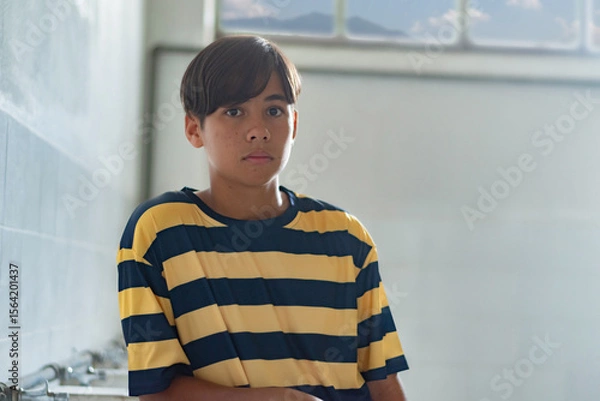 Fototapeta A teenage boy stands quietly indoors with a serious expression, wearing a striped shirt. Natural light comes through the window, adding a soft atmosphere.