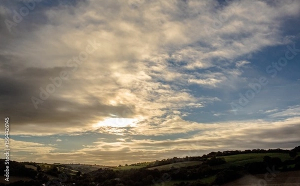Fototapeta Sunlight is filtering through dramatic sky over rolling green hills and farmland in flat design