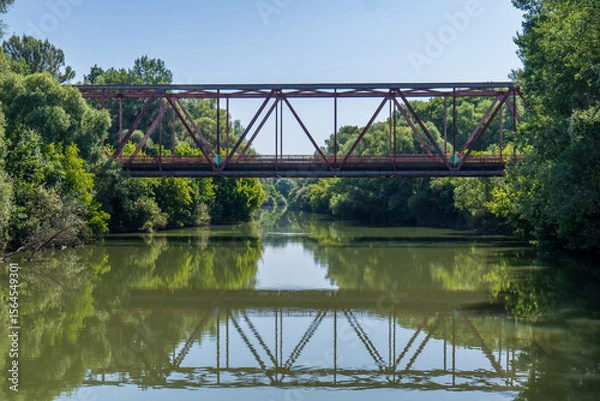 Fototapeta Metal bridge over the Bodrog river in Viničky village, Slovakia. Green trees on both sides and calm water reflecting the bridge structure on a sunny summer day.