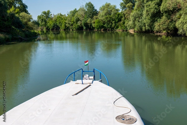 Fototapeta Boat on Bodrog river with Hungarian flag, green trees, calm water, blue sky and mooring rope on the deck. Summer river landscape between Slovakia and Hungary.