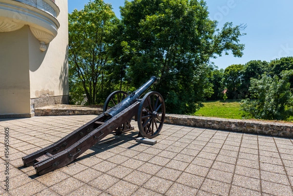 Fototapeta Old black cannon on wooden wheels placed on a stone terrace near Sárospatak Castle, Hungary, with green trees in the background on a sunny day.