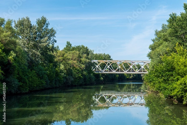 Fototapeta Railway bridge over Bodrog river surrounded by green trees. Bridge and trees reflected in calm water on a sunny summer day.
