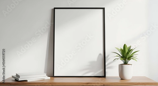 Fototapeta A blank framed poster stands on a wooden table next to a potted plant, with sunlight casting shadows on the wall.