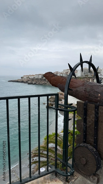 Fototapeta Nerja, Spain - June 02, 2025: Nerja, balcony of Europe, beach, old town, historical center, tourism. Scenic coastal view from a black iron railing overlooking the ocean and distant buildings