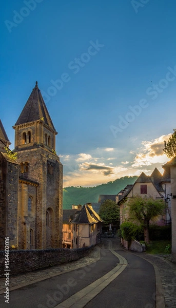 Fototapeta Winding cobblestone street through the historic French village of Conques at golden hour with the evening light illuminating the tower of the Abbey of Sainte-Foy and medieval houses under a blue sky,