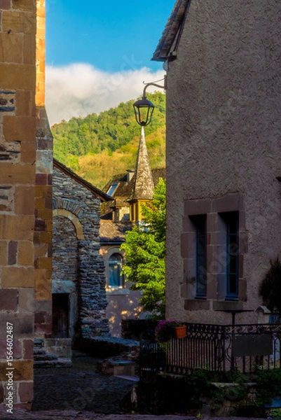 Fototapeta A hidden alleyway off a medieval street with the golden light of sunset illuminating the spire of a stone church tower and the green hills in the background in Conques, France.