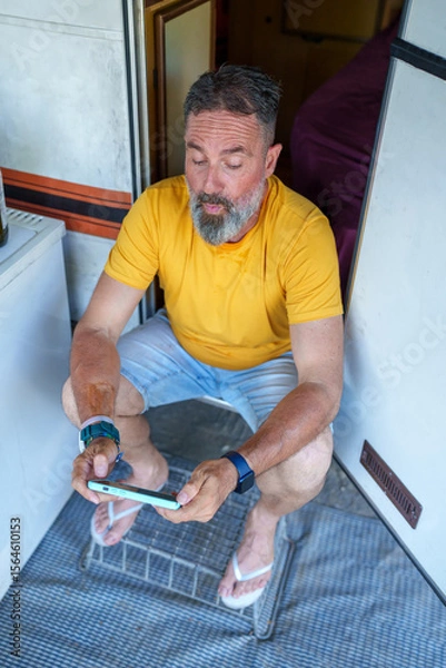 Fototapeta Bearded traveler relaxing inside camper van, playing mobile game during summer road trip with casual smartphone engagement