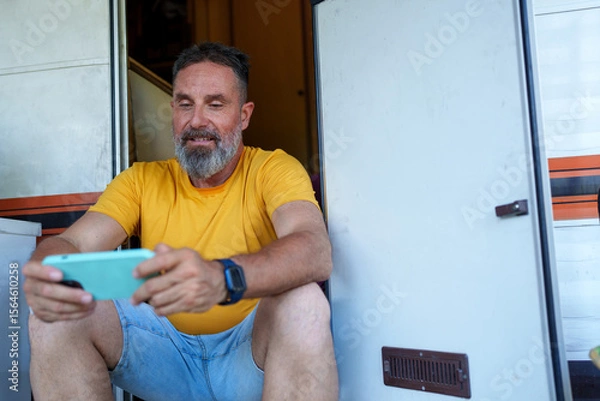 Fototapeta Happy camper playing mobile games on his phone while sitting on the steps of his recreational vehicle
