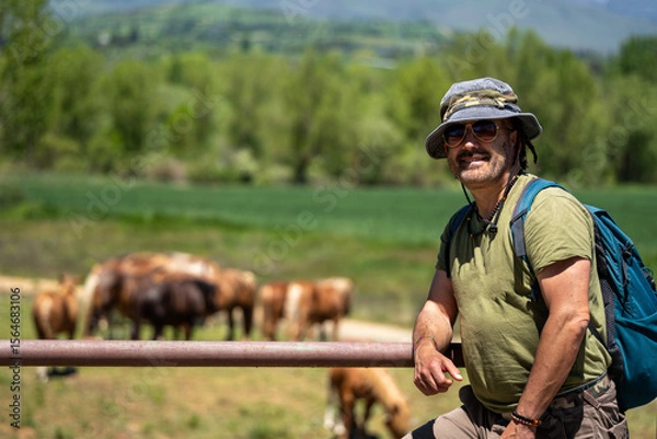 Fototapeta Caucasian hiker with backpack and hat enjoying mountain landscape with horses grazing in the background