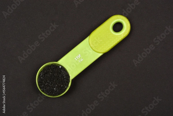 Obraz A vibrant green 1 teaspoon (5 ml) measuring spoon filled with black Hawaiian salt is centrally positioned on a dark background