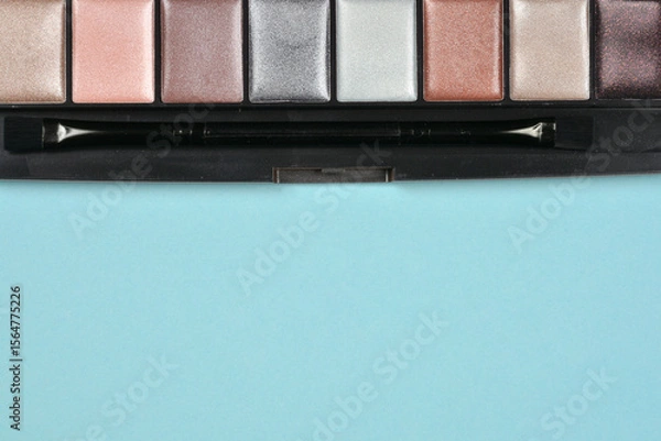Obraz Shimmering eyeshadow palette with various metallic shades of silver, pink, and brown, including a small applicator brush, on a light blue background