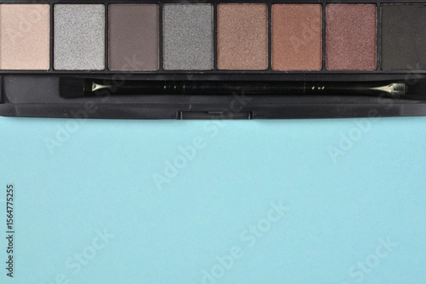 Obraz Dark eyeshadow palette with various shades of brown and gray, including a small applicator brush, on a light blue background.