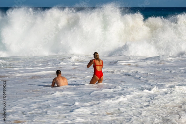 Fototapeta Couple standing on sandy beach, gazing at a powerful wave crashing under a moody sky. Moment of awe, connection, and raw nature. Adventure captured by the ocean’s edge.