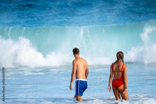 Fototapeta Couple standing on sandy beach, gazing at a powerful wave crashing under a moody sky. Moment of awe, connection, and raw nature. Adventure captured by the ocean’s edge.