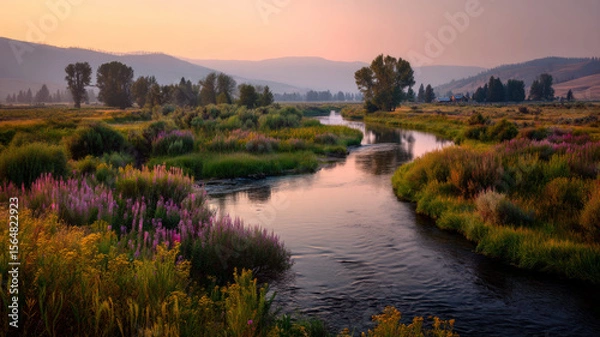 Fototapeta A river winds through a landscape glowing with sunset light, its surface reflecting golden hues.
The banks are lined with wildflowers, ferns, and willows; distant hills fade near a farmhouse.