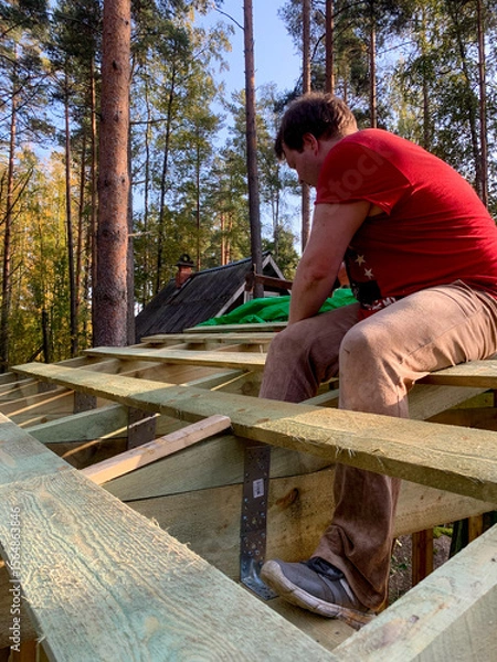 Fototapeta A Caucasian man in a vibrant red shirt builds dreamlike forest cabin, capturing rustic nostalgia, Arbor Day spirit