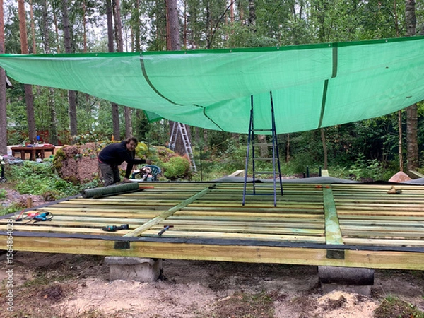 Fototapeta Crafting a rustic forest sanctuary, a focused Middle-Eastern man assembles a deck under a verdant shade tarp, celebrating International Building Safety Week