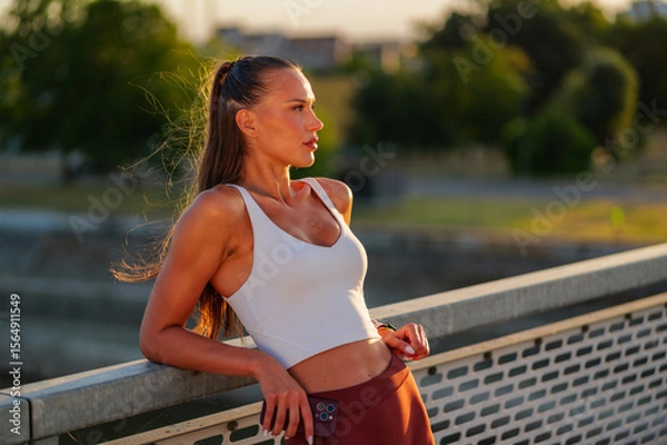 Fototapeta Fit young woman in activewear leaning on a bridge railing, enjoying a serene sunset. She embodies a healthy lifestyle and mindfulness in an urban park setting