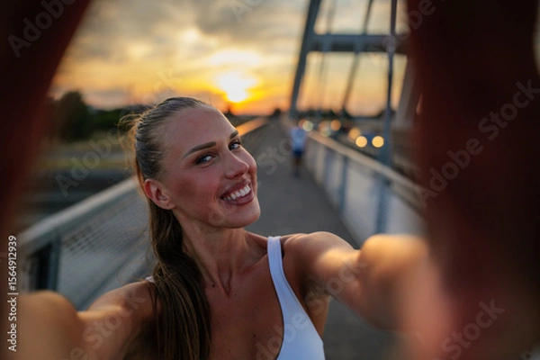 Fototapeta Smiling sportswoman taking a selfie on a bridge during sunset, enjoying the warm golden hour light after a refreshing workout, celebrating an active and healthy lifestyle