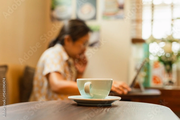 Fototapeta Selected focus cup of coffee on table with woman working on laptop in blur background.