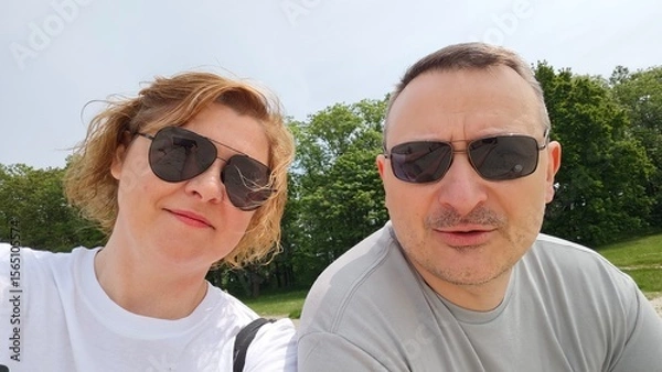 Fototapeta Happy middle-aged couple wearing sunglasses taking a selfie outdoors on a sunny day, smiling together in a green park environment