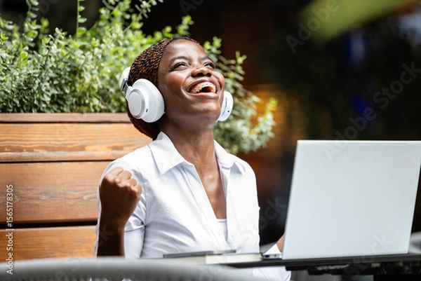 Obraz Elegant black African woman in business suit working on laptop computer and celebrating success in cafe bar.