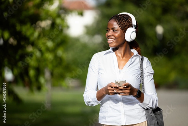 Obraz Smiling black African woman walking through the park and using smart phone.