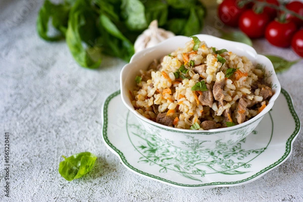 Fototapeta A hearty lunch: pearl barley with meat and vegetables on a beautiful white plate on a light background. Pearl barley with pork for dinner on a beautiful plate. Close-up