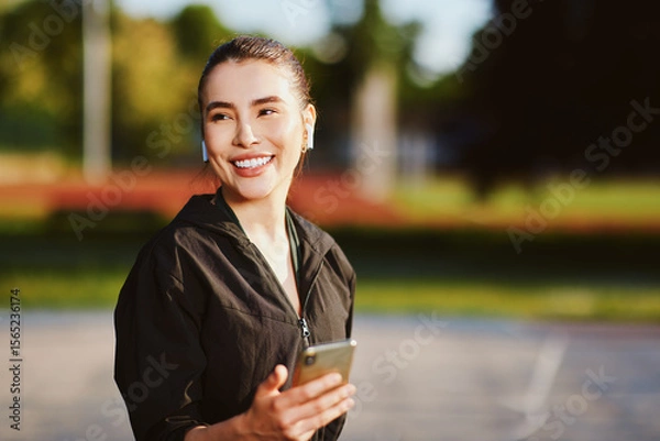 Obraz Smiling sporty woman listening music using wireless earbuds and holding smartphone in a park