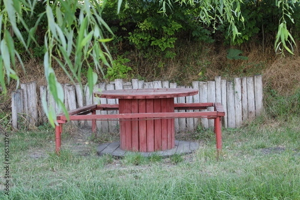 Obraz A red wooden picnic table surrounded by greenery and wooden fencing.