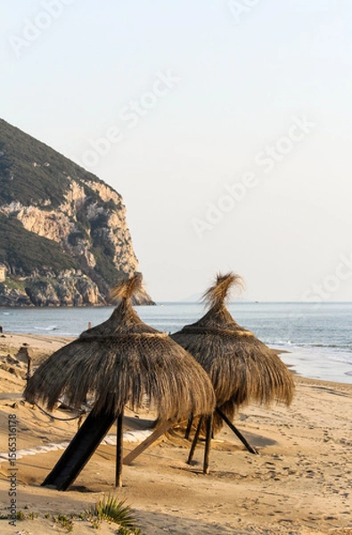 Fototapeta Two straw beach umbrellas on a sandy shore with rocky cliff and calm sea in the background at sunset. Peaceful coastal scenery and natural beach setting. Outdoor vacation and summer atmosphere.