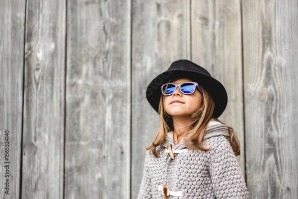Fototapeta Confident child girl wearing a black hat and blue sunglasses poses in front of a rustic wooden wall. Her style and expression convey individuality and self-confidence. Generation alpha concept.