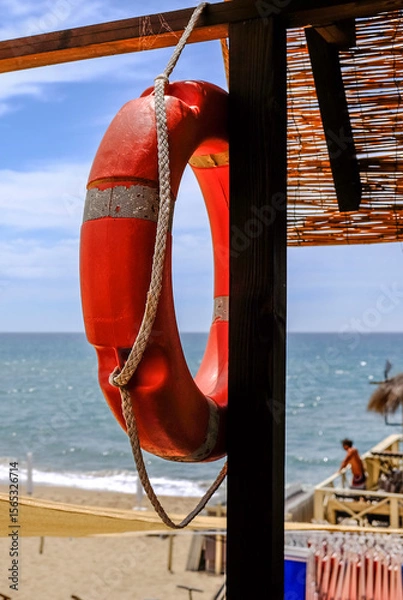 Fototapeta Red lifebuoy hanging on a wooden beach hut with the sea and sandy shore in the background. Bright summer day with strong sunlight and beach atmosphere. Safety and vacation concept.