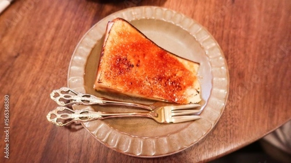 Fototapeta close up of a slice of baked basque cheese cake looks like creme brulee custard cake with caramelized top with a glossy, crackled surface on an elegant plate with two ornate silver forks