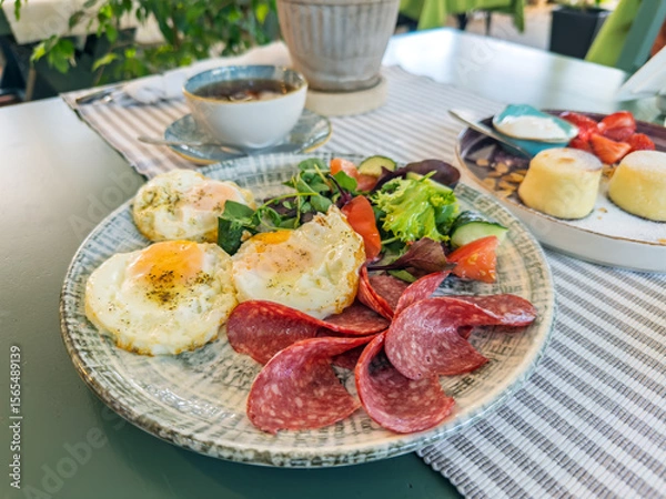 Obraz A delicious and hearty breakfast plate with fried eggs, salami, and fresh vegetable salad. A complete morning meal served in a cafe