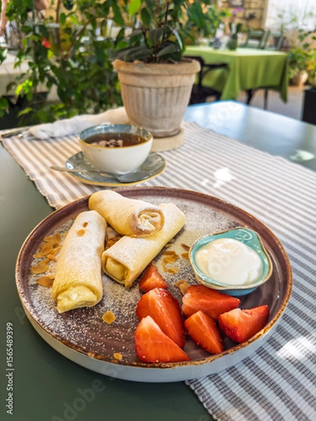 Obraz A delicious sweet breakfast with cheese-filled crepes (blini), fresh strawberries, and sour cream, served with tea in a cozy cafe.A healthy and tasty brunch in a restaurant.