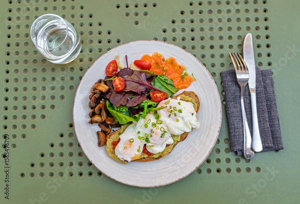 Obraz Gourmet breakfast plate with poached eggs on avocado toast, smoked salmon, and fresh salad.Top view.Healthy and balanced brunch with avocado toast, poached eggs, smoked salmon, and side of mushrooms