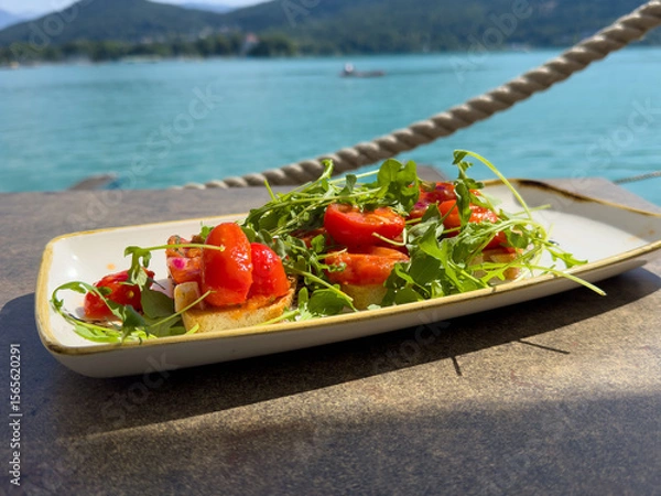 Fototapeta Fresh bruschetta appetizer with arugula and cherry tomatoes by scenic lake view