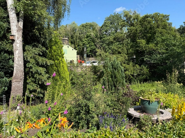 Fototapeta “Beneath a clear blue hush, tall trees and bright blossoms sway in a sunlit garden. Amid the green, a house of jade quietly nestles, half-dreamt through the veil of foliage on, Goit Stock Lane, Harden