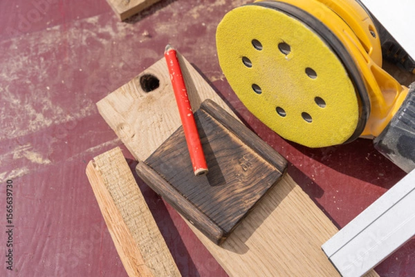 Fototapeta Woodworking tools and materials laid out on a workbench, including a yellow orbital sander, red carpenter's pencil, various wood pieces, and a metal square ruler.