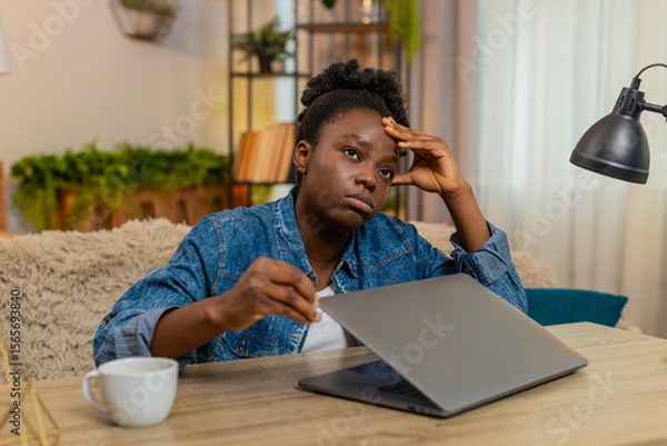 Fototapeta African American young woman closes laptop with visible stress after a hard day of online learning while sitting at table on home sofa. Black girl holds head, overwhelmed by digital study fatigue.