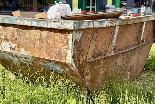 Obraz An Old and Rusty Dump Container That is Surrounded by Lush, Overgrown Grass and Weeds. A weathered metal dumpster overflowing with various debris, positioned against a backdrop of lush tall grass.