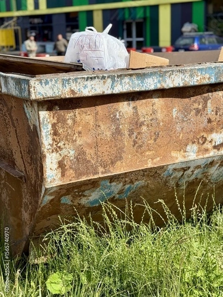 Obraz An Old and Rusty Dump Container That is Surrounded by Lush, Overgrown Grass and Weeds. A weathered metal dumpster overflowing with various debris, positioned against a backdrop of lush tall grass.