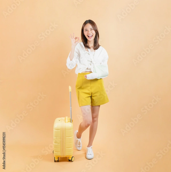 Fototapeta Asian woman broken arm wrapped and shows ok sign with suitcase isolated on beige color background, Travel insurance concepts.
