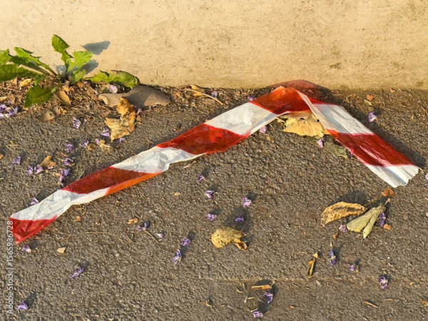 Fototapeta Red and white caution tape on roadside with fallen leaves and flowers