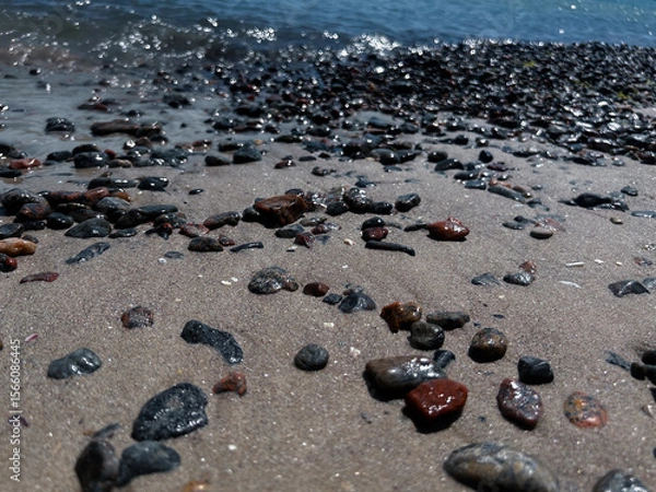 Fototapeta Close-up of wet pebbles scattered on a sandy beach in Odesa, Ukraine, with gentle sea waves sparkling in the background. The sunlit stones create a natural texture and coastal atmosphere, perfect for