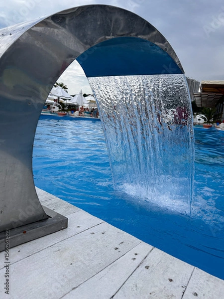 Fototapeta Modern outdoor swimming pool with waterfall fountain, people relaxing in background, summer leisure concept.