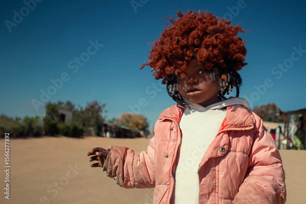 Fototapeta A young African girl with dark skin, wearing a vibrant orange wig and pink jacket. A real moment of playful expression, captured in natural light. Perfect for diversity, childhood, and lifestyle theme