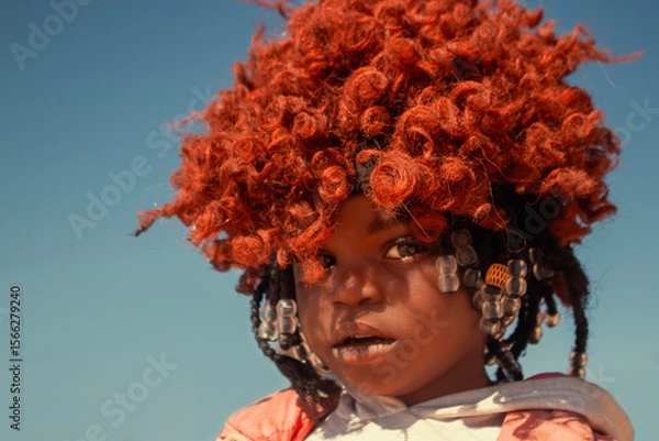 Fototapeta A young African girl with dark skin, wearing a vibrant orange wig and pink jacket. A real moment of playful expression, captured in natural light. Perfect for diversity, childhood, and lifestyle theme
