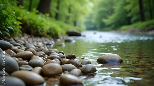 Fototapeta A high-resolution photo of smooth pebbles arranged naturally along the edge of a clear stream, water gently flowing and reflecting nearby greenery -